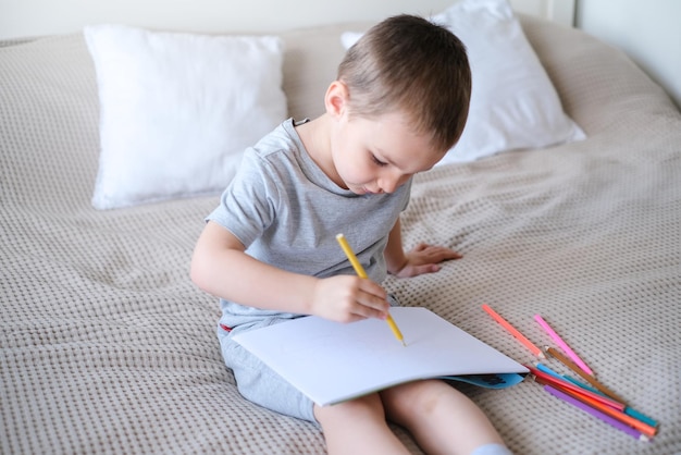 A small caucasian boy in a gray Tshirt and shorts on the bed draws with colored pencils in a sketchb