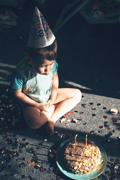 Small caucasian boy with party hat is blowing the birthday lights sitting on the floor near confetti