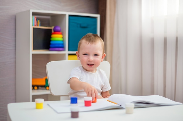 A small cheerful boy is sitting at a table and drawing with paints