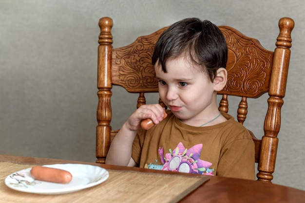 A small child boy eats a sausage at the table