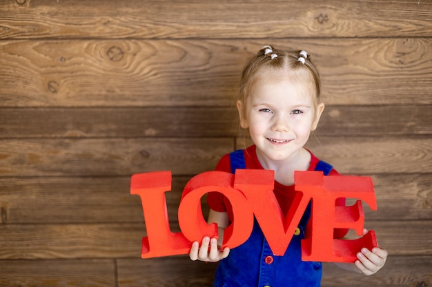 A small child girl with a red inscription Love in her hands smiles or laughs against a wooden wall the concept of Valentine's day an empty space for text