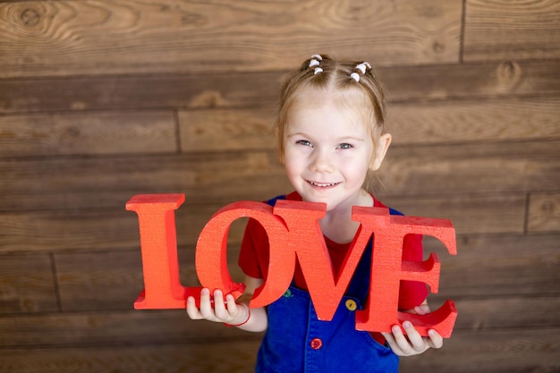 A small child girl with a red inscription Love in her hands smiles or laughs against a wooden wall the concept of Valentines day an empty space for text