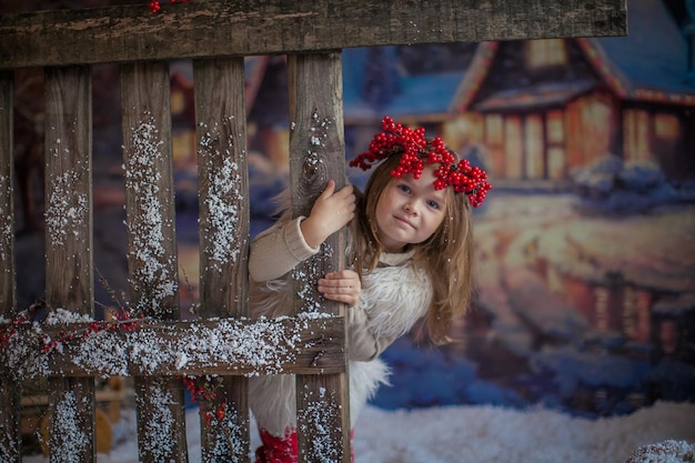 A small child girl with a wreath of rowan on her head near a natural wooden fence