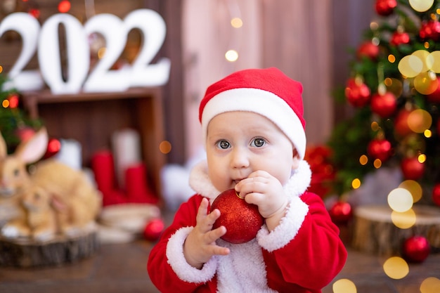 A small child is sitting with Christmas trees. A large portrait. A baby in a red Santa Claus costume with Christmas tree toys. Happy Holidays, New Year. Christmas Time