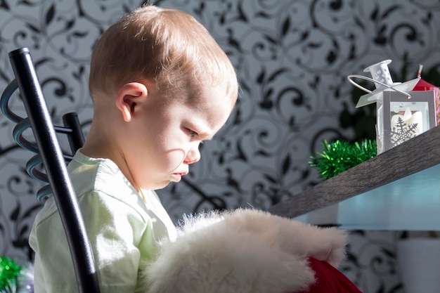 A small child in a Santa Claus hat and red mittens with Christmas gifts is sitting at the New Year's table