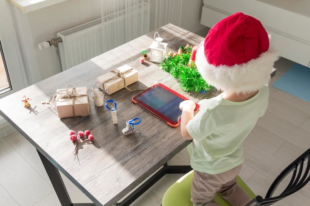 A small child in a Santa Claus hat with Christmas gifts is sitting at a New Year's table with a tablet in his hands