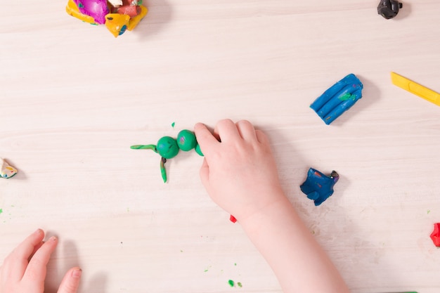 A small child sculpts a caterpillar from green plasticine on a wooden table