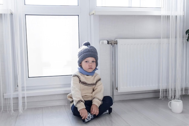 A small child in a sweater and a gray hat is kneeling near a heater with a thermostat