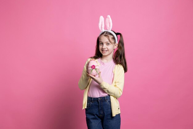 Small child with bunny ears holding a painted pink egg in front of camera