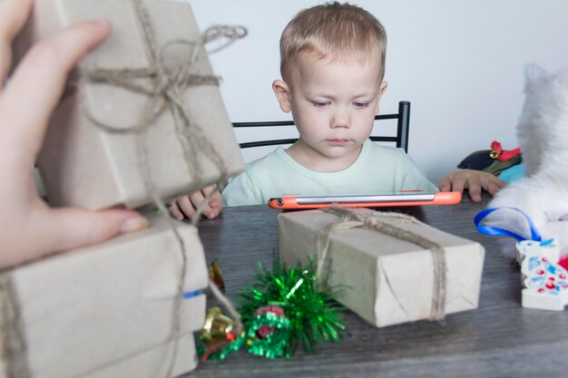 A small child with Christmas gifts is sitting at a New Year's wooden table with a tablet in his hands