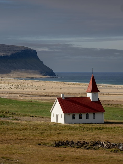 A small church with a red roof sits in a field next to a cliff.