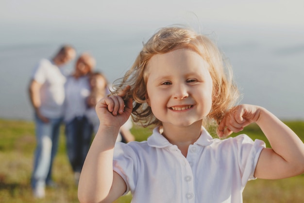 A small curly-haired girl in white looks into the frame and smiles against the background of her parents in the summer in nature