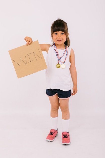 Small dark-haired girl with sports champion medals, holding a sign that reads: 'win'. Sport and victory concept