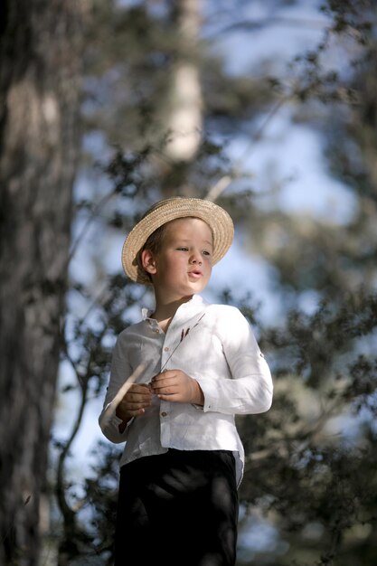 Small farm boy smiling and looking at the camera stands in forest Cute kid boy in a straw hat