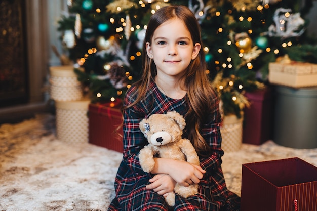 Small female child with appealing appearance, holds teddy bear, recieves present from parents, sits near Christmas tree