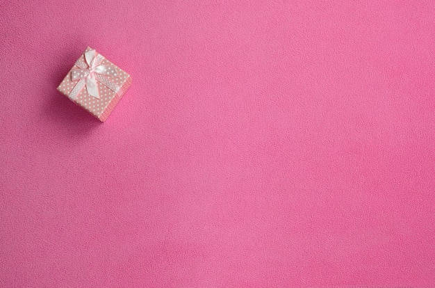 A small gift box in pink with a small bow lies on a blanket of soft and furry light pink fleece fabric.