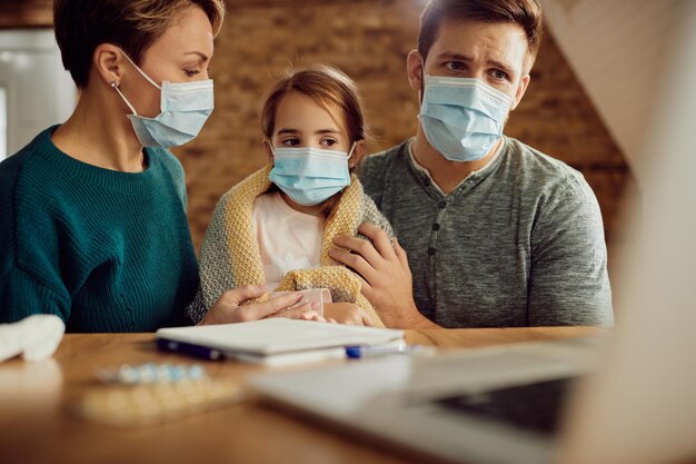 Small girl and her worried parents wearing face masks while using laptop at home