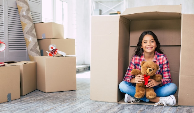 A small girl holding a teddy bear is hiding in a cardboard box, probably playing hide and seek with her parents in their spacious apartment