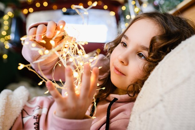 Small girl illuminated face, holding garland warm yellow lights at Christmas and New year background