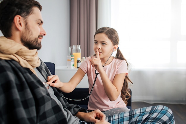 Small girl listen to dad's breathing through stethoscope. She hold finger on lips and look at him. Sick young man covered with blanket and scarf.