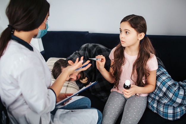 Small girl look at female doctor. She hold thermometer and bottle of syrup. Girl talk to doctor. Sick young man sleeping on couch. He covered with blanket.