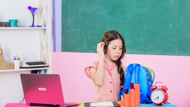 Small girl pupil with computer Use new technology back to school Online education homeschooling concept Good morning study online Lesson alarm girl in class with clock wait for day end