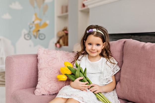 A small girl in a white dress with a bouquet of tulips is sitting on the sofa in the room.