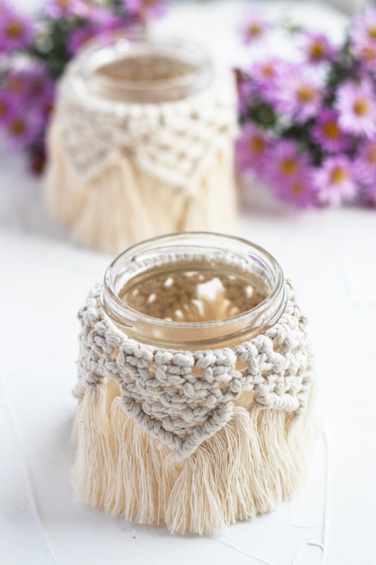 Small glass vase, jar, candleholder with macrame cover. Autumn flowers blurred on background. Boho