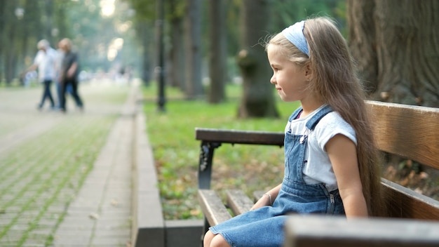 Small happy child girl sitting on a bench resting in summer park.