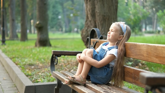 Small happy child girl sitting on a bench resting in summer park