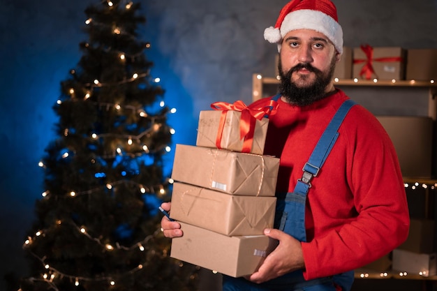 Photo small and medium business startup concept a young startup businessman wearing a santa claus hat and work overalls takes a stand in a home office holding a lot of boxes in his hands