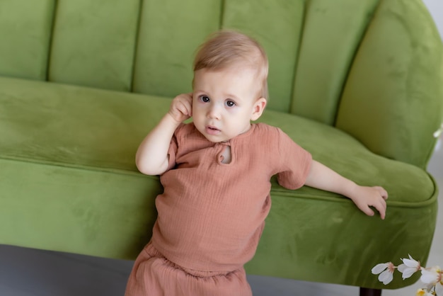 Small newborn boy in a beige suit stands near a green sofa surrounded by beautiful flowers Looking at the camera