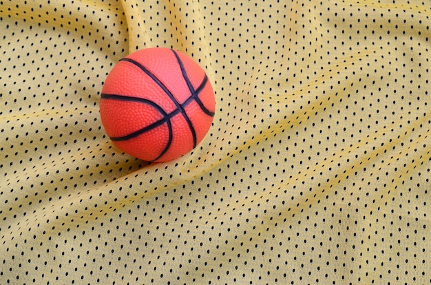 Small orange rubber basketball lies on a yellow sport jersey clothing fabric texture