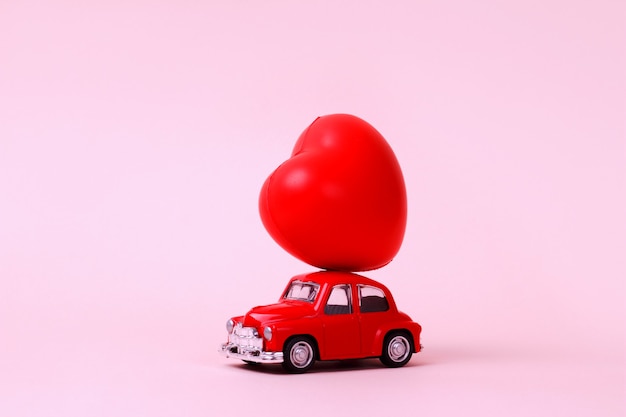 Small red retro toy car with heart on the roof on pink Valentines Day present delivery concept