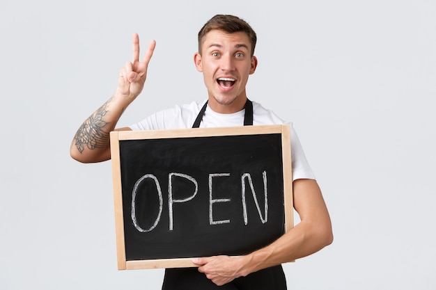 Small retail business owners, cafe and restaurant employees concept. Upbeat handsome waiter, salesman in black apron, showing peace sign, greeting guests as holding open sign