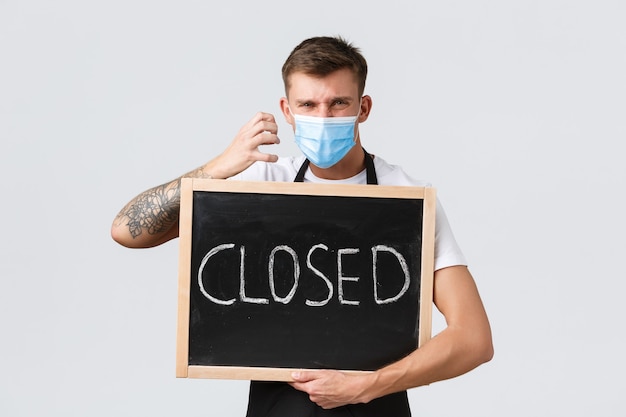 Small retail business owners, covid-19 and social distancing concept. Outraged and disappointed businessman, salesman in medical mask shaking fist angry and showing closed sign