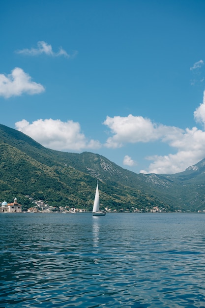 A small sailing yacht floats on the sea by the green mountains, blue sky and clouds