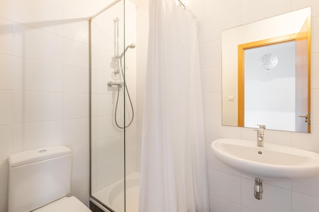 Small simple bright bathroom with shower area enclosed by glass partition and screen Square mirror above washbasin shows front door to bedroom There is white toilet against wall