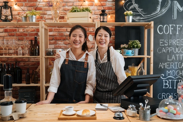 Small startup business owner concept. two successful young baristas women standing in bar counter in cafe. happy coffeehouse waitresses in aprons smiling confidently to camera in coffee shop.