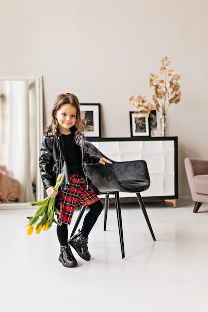 A small stylish girl in a black leather jacket and a plaid skirt with a bouquet of tulips is standing in the room.