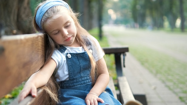 Small tired child girl sitting on a bench with closed eyes resting in summer park.