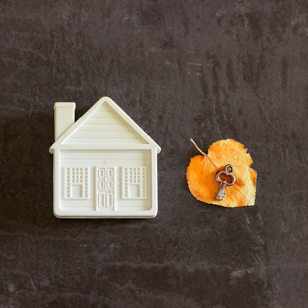 Next to a small toy house is a key on a yellow autumn leaf. 