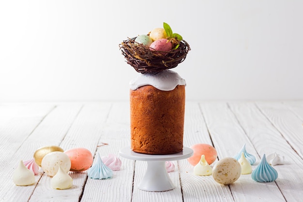 Small traditional Paska bread on cake stand glazed and decorated with nest Few colorful eggs in pastel colours and meringue biscuits placed around Minimalistic style Copy space