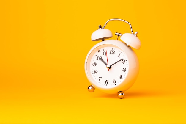 Small white alarm clock, black numbers, set the time placed on a table. Clock on isolated yellow background.