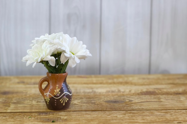 Small white fresh daisies in a brown clay vase on a light wooden background closeup