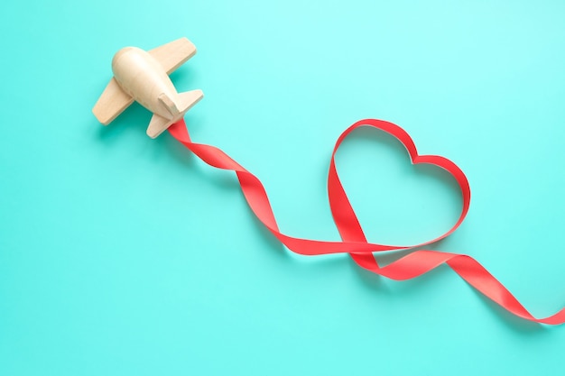 A small wooden toy airplane carries Valentine's elements. Vapor trail of sequins in the shape of heart, and red ribbon in the shape of heart. Valentine's day