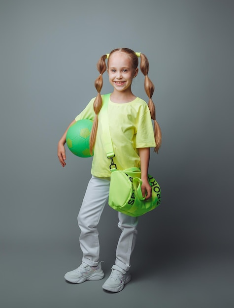 A small young girl with a green sword in her hand smiles at the camera isolated on a gray background a little athlete with a ball children's sports