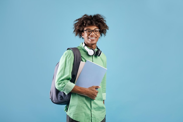 Smart african american student guy with backpack and notebooks smiling at camera blue studio