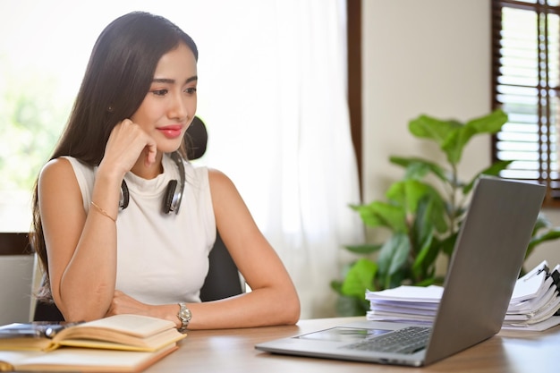 Smart Asian female boss looking at laptop screen hand on chin thinking about new strategy