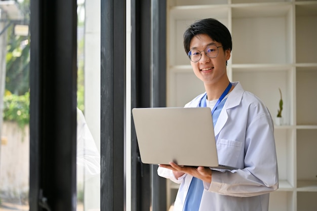 Smart Asian male medical student in white gown stands in a hospital corridor with his laptop